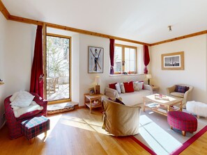 Ein geräumiges Wohnzimmer mit einer Couch, Sesseln, einem Couchtisch und einem Kamin. Ein großes Fenster sorgt für natürliches Licht und bietet einen Blick ins Freie.