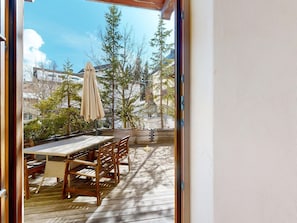 Eine Holzterrasse mit Tisch, Stühlen und Sonnenschirm, mit Blick auf Bäume und Gebäude.