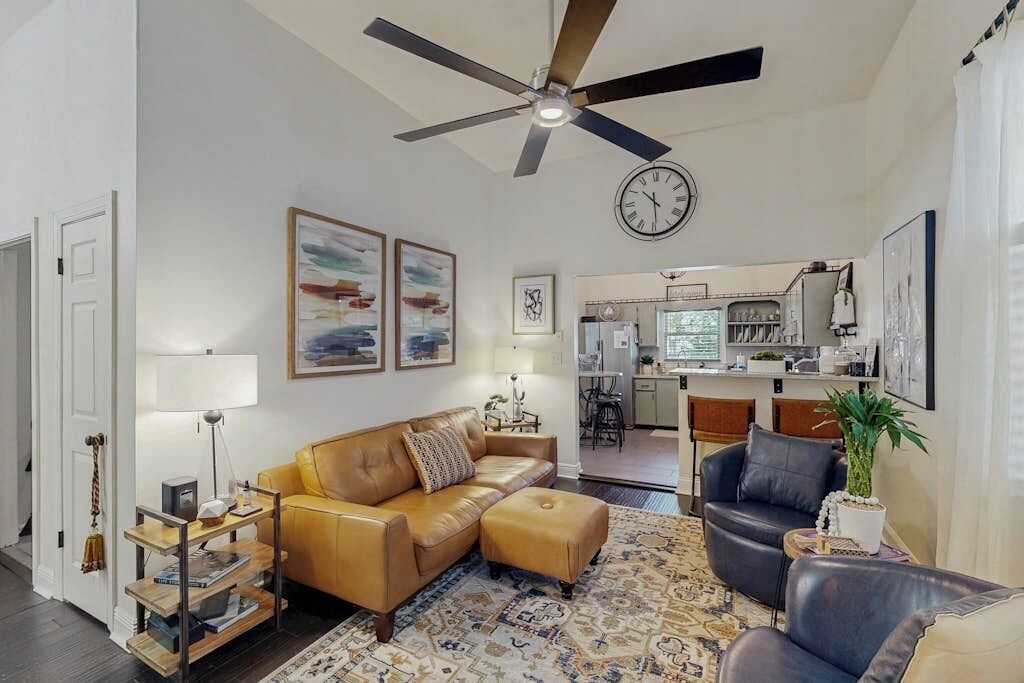 Un salon comprenant un canapé, un pouf et des fauteuils, un ventilateur de plafond, une horloge et des œuvres d’art accrochées aux murs.