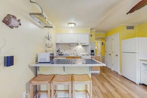 A compact kitchen with white cabinets, a microwave, and a refrigerator. There are wooden stools at the counter and a fish decoration on the wall. - Waterfront island getaway with balcony, pool, tennis, & easy beach access (Tybee Island)