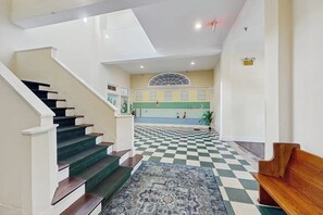 Um hall de hotel com piso quadriculado, uma escadaria com degraus escuros e uma mesa de recepção.
