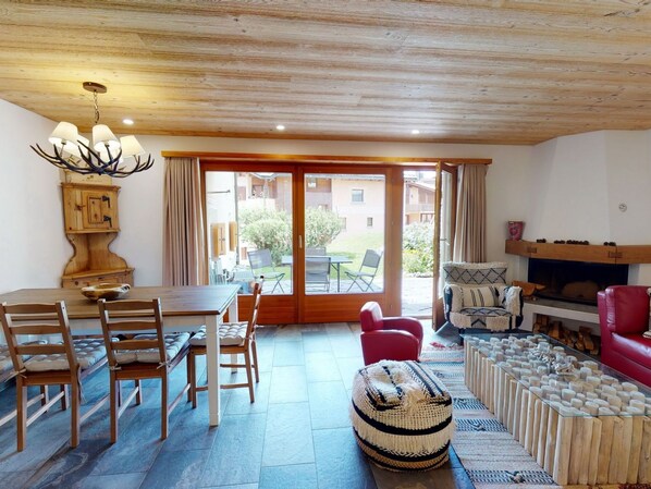 Una habitació acollidora amb sostre de fusta, una taula de menjador amb cadires, una llar de foc i una finestra gran amb cortines.
