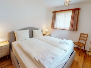 Uma cama bem arrumada com lençóis brancos e uma toalha dobrada, uma mesa de cabeceira com abajur, uma cadeira de madeira e uma janela com cortinas.