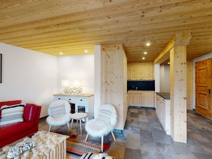 Ein gemütliches Interieur mit einem roten Sofa, zwei weißen Sesseln, einem kleinen Tisch und einer Küchenzeile mit Holzschränken und einer Arbeitsplatte.