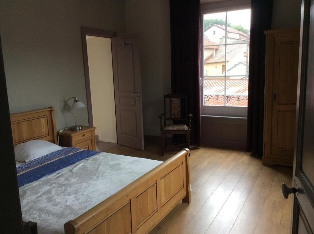 Une chambre à coucher avec un lit en bois, une table de chevet, une chaise, une fenêtre donnant sur des immeubles et une porte.