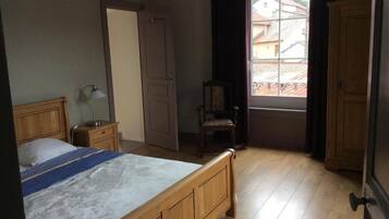Une chambre à coucher avec un lit en bois, une table de chevet, une chaise, une fenêtre donnant sur des immeubles et une porte.