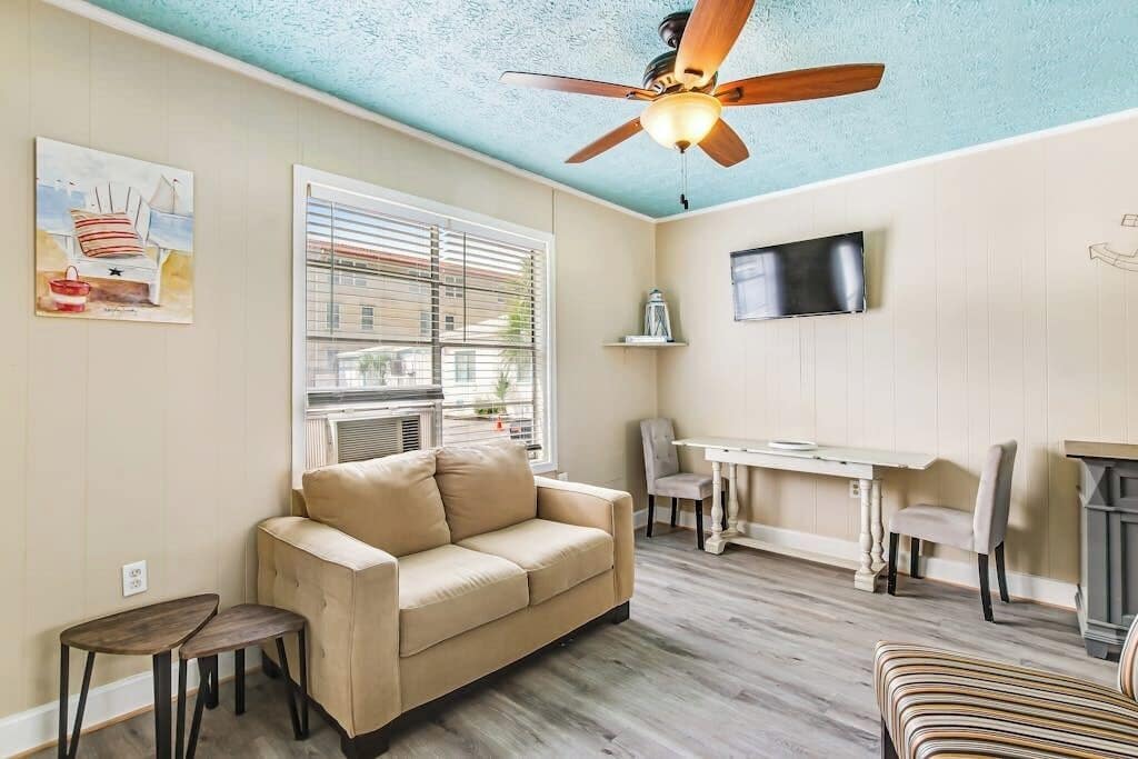 A living room with a beige sofa, a small table, a ceiling fan, and a TV mounted on the wall.