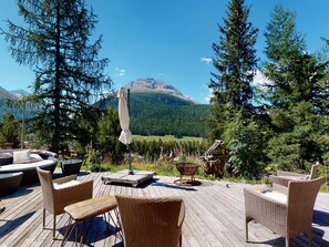 Eine Holzterrasse mit Korbmöbeln, einem Tisch und einem Sonnenschirm, mit Blick auf eine gebirgige Landschaft.