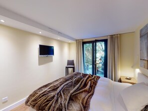 Ein Schlafzimmer mit einem Bett, einem an der Wand montierten Fernseher, einem Sessel und einem Fenster mit Vorhängen.