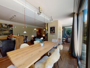 Ein moderner Essbereich mit einem Holztisch, weißen, flauschigen Stühlen, Pendelleuchten und Blick durch große Fenster.