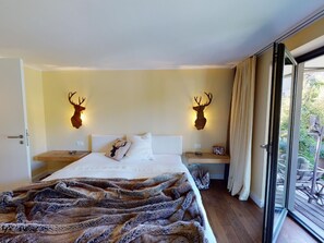 Ein Schlafzimmer mit einem Bett, zwei an der Wand befestigten Hirschgeweihen, einem Fenster mit Vorhängen und Blick ins Grüne.