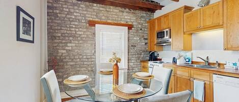 A dining area with a glass table and chairs, a brick wall, wooden beams, and a kitchen with wooden cabinets and appliances.