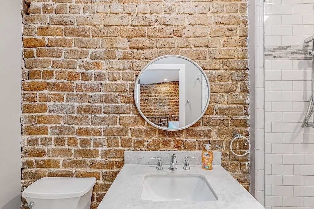 Ein Badezimmer mit Backsteinwand, einem runden Spiegel, einem weißen Waschbecken und einer Toilette.