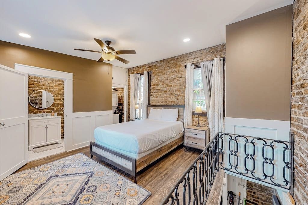Une chambre à coucher avec un lit, des tables de chevet, un ventilateur de plafond, une fenêtre avec des rideaux et un mur en briques.
