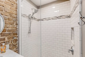 A shower with a glass door, white tiled walls, and a brick accent wall. - Atmospheric carriage house steps from Forsyth Park, great for long stays (Savannah)
