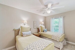 A bedroom with two beds, a ceiling fan, bedside lamps, a small round table, and a window with curtains. - Seaside serenity home with beach access, washer/dryer, & dog-friendly (Tybee Island)