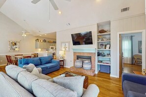 A living room with a fireplace, a television, and a dining area with a kitchen in the background. - Seaside serenity home with beach access, washer/dryer, & dog-friendly (Tybee Island)