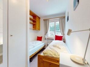 Una habitación pequeña y bien iluminada con dos camas individuales, un armario de madera, una ventana con cortinas y una lámpara de noche.