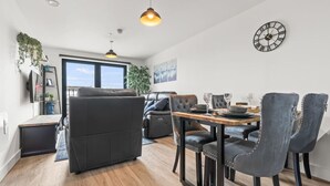 A modern living room with a sofa, dining table, and chairs, a large window, and a wall clock. - Central Liverpool 2 Bedroom Apartment, Large, Modern, WIFI, Sleeps 4 (Liverpool)