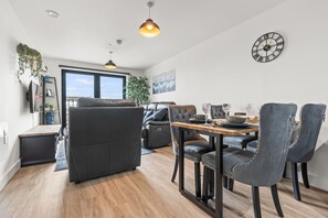 Ein modernes Wohnzimmer mit einer Couch, einem Esstisch, Stühlen, einem großen Fenster und einer Wanduhr.