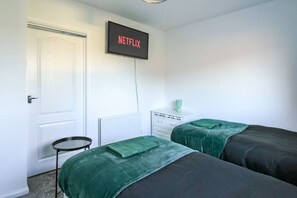 Une chambre avec deux lits, une petite table, une commode, un radiateur et un téléviseur fixé au mur qui affiche l’écran d’accueil de « NETFLIX ».