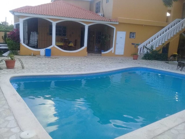 Una piscina con solárium empedrado, un porche cubierto y una escalera que lleva a una puerta.