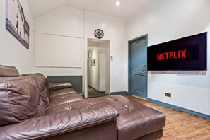 Une pièce avec un canapé en cuir marron, un téléviseur fixé au mur qui affiche le logo « NETFLIX », et une porte dotée d’un cadre bleu.