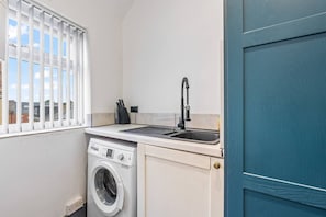 A compact kitchen with a washing machine, a sink, and a window with blinds. - Stylish Homely Flat|Parking|4 Beds|6 Guests (Sheffield)