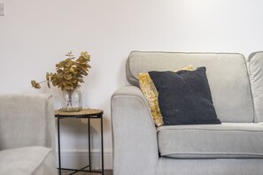 Ein Wohnzimmer mit grauem Sofa, einem kleinen schwarzen Beistelltisch und einer Vase mit getrockneten Blumen.
