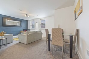 A modern living room with a sofa, a glass dining table, and chairs. There is a large mirror on the wall and a window with blinds. - City Centre Derby | Modern 2-Bed Flat (Derby)