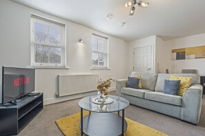 A modern living room with a television, a glass coffee table, a grey sofa with patterned cushions, and a radiator. - City Centre Derby | Modern 2-Bed Flat (Derby)