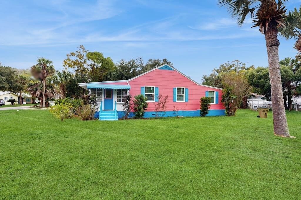 A single-story house with a red and blue exterior, surrounded by a well-maintained lawn and palm trees.