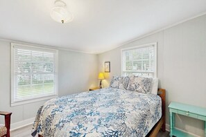 A bedroom with a bed, two windows with blinds, a nightstand with a lamp, and a small table. - Beachy dog-friendly home, blocks to the restaurants, fishing pier & beach (Tybee Island)