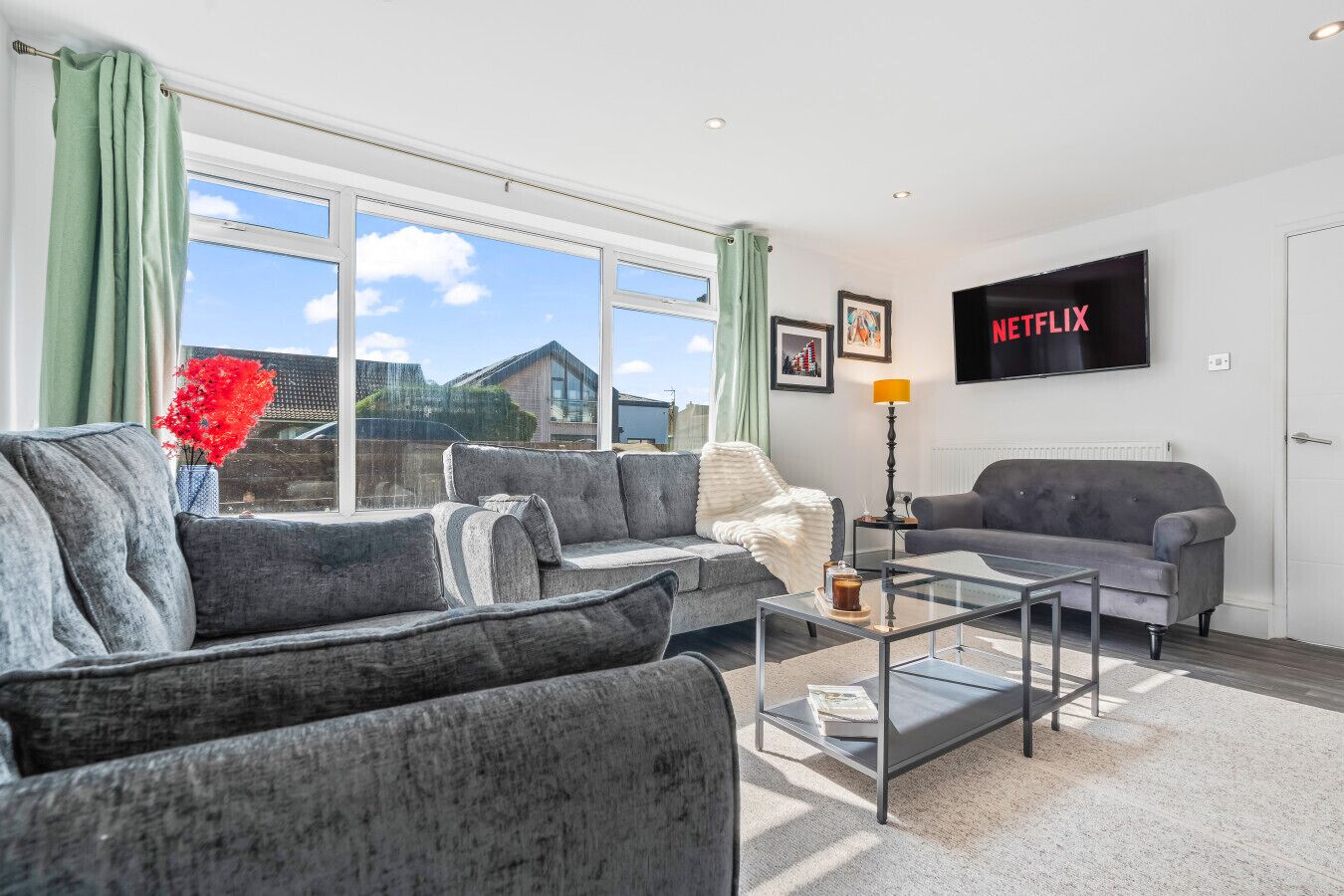 Een moderne woonkamer met grijze banken, een glazen salontafel en een groot raam met gordijnen. Een flatscreen televisie hangt aan de muur.