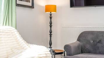 Una habitación con un sillón gris, una lámpara de pie, un cuadro enmarcado y un televisor en la pared.