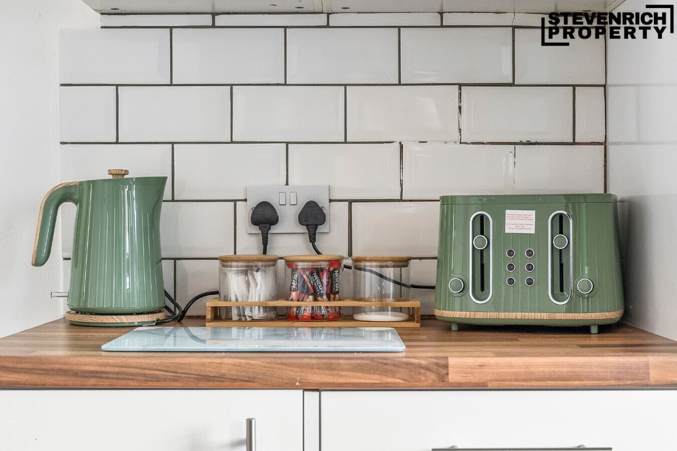 A kitchen with a white tiled backsplash, a green kettle, a toaster, and jars on a wooden board.