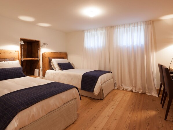 Quarto de hotel com duas camas, cabeceiras de madeira, uma escrivaninha e uma janela grande com cortinas.