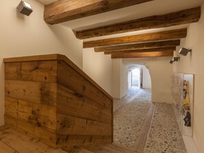 Uma escada de madeira leva a um corredor com chão de pedra e uma porta no final.