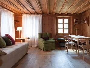 A cozy cabin interior with a wooden ceiling, a sofa with cushions, a green armchair, a small table with a lamp, and a desk with chairs. - Chalet Ches'Ota by Interhome (La Punt-Chamues-ch)