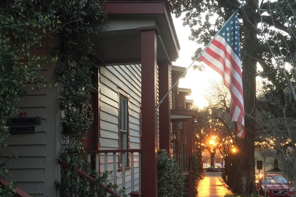 A residential building with a porch, an American flag, and a street with parked cars.