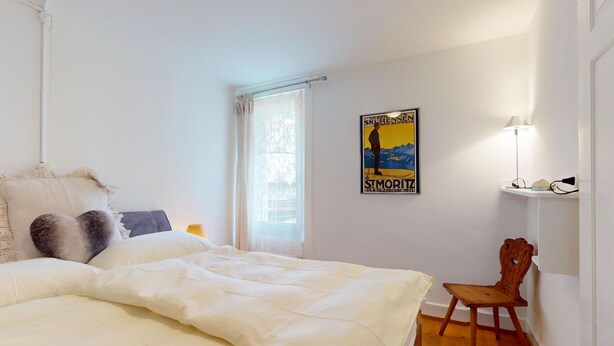Un dormitorio con una cama grande, una silla, una ventana con cortinas, una lámpara de pared y un cartel.