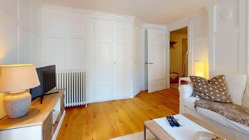 A living room with a sofa, a coffee table, a TV, and a lamp. There is a radiator and a door leading to another room.