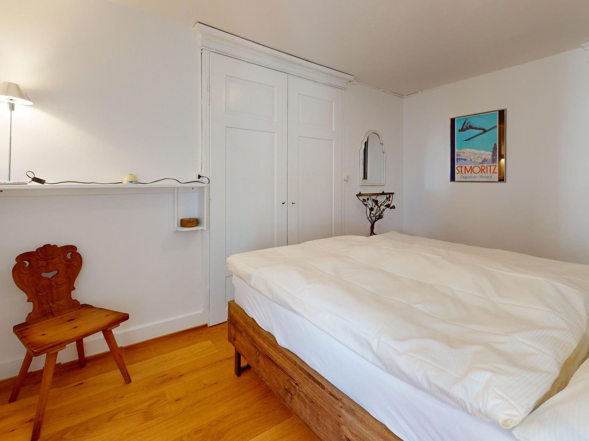 Slaapkamer met een houten bed, een stoel, een nachtkastje, een wandlamp, een spiegel en een lijst met een afbeelding.