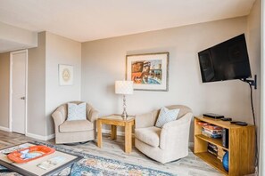 A living room with two sofas, a coffee table, a TV mounted on the wall, and a bookshelf with books and a board game. - Oceanfront gem with spacious deck, direct beach access, & high-speed WiFi (Tybee Island)