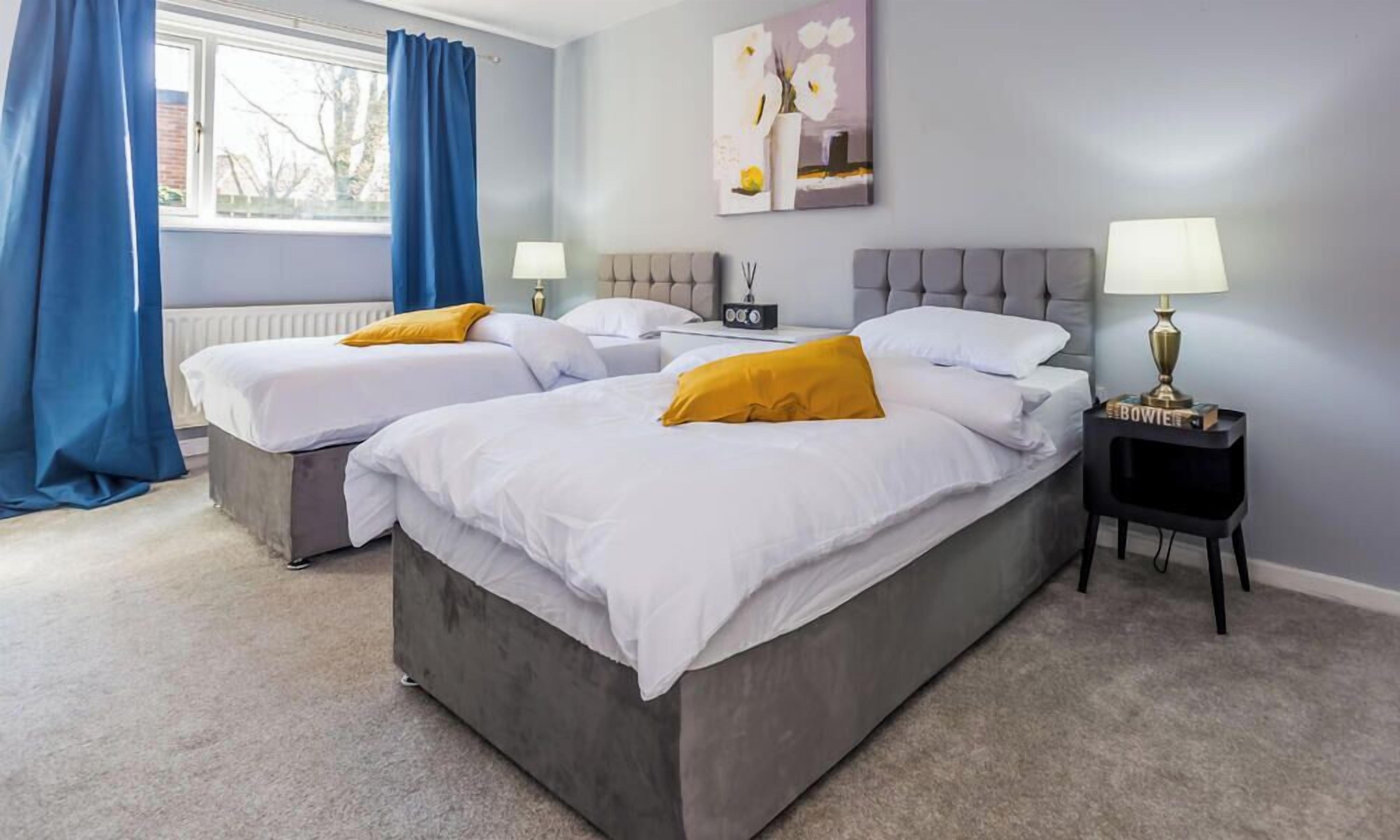 Ein Hotelzimmer mit zwei Betten, einem Fenster mit Vorhängen, einem Nachttisch mit Lampe und einem Bild an der Wand.