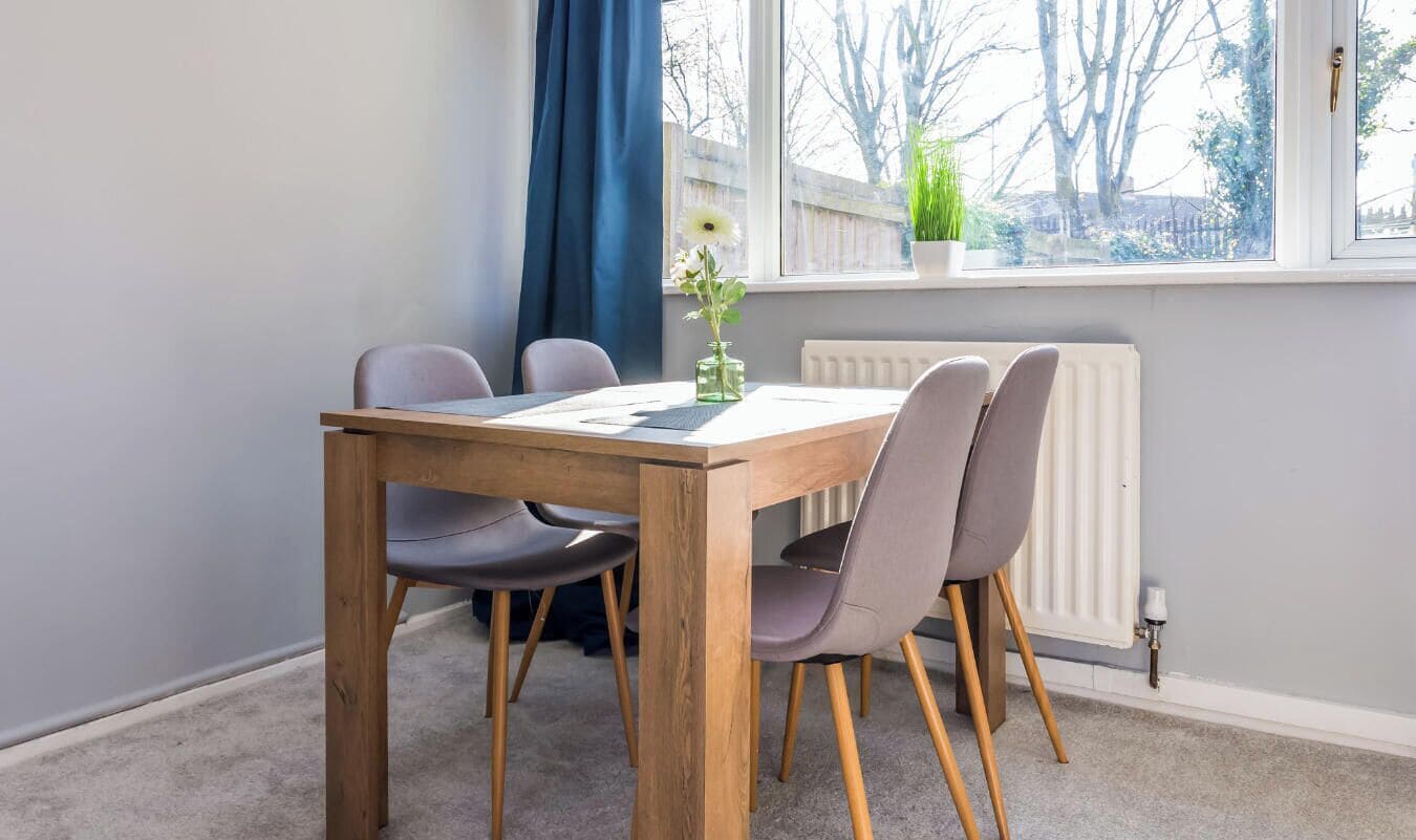 A dining area with a wooden table and four chairs, a window with a view of trees, and a radiator.