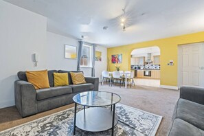 Ein modernes Wohnzimmer mit einem grauen Sofa, einem Glastisch und einem Essbereich mit weißen Stühlen.