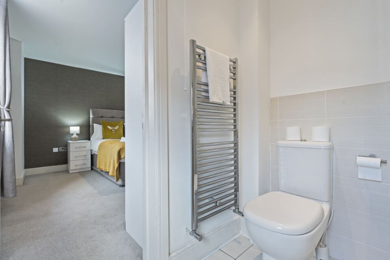 Een moderne badkamer met toilet, een handdoekhouder, en een bed in de aangrenzende kamer.