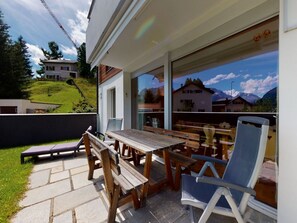 Um pátio externo com mesa e bancos de madeira, uma cadeira e vista para uma paisagem montanhosa através de grandes janelas.