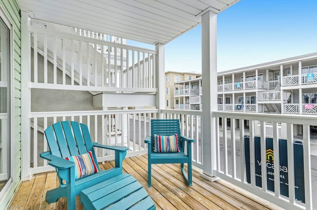 A covered outdoor area with two blue Adirondack chairs, white railings, and a view of a multi-story building.